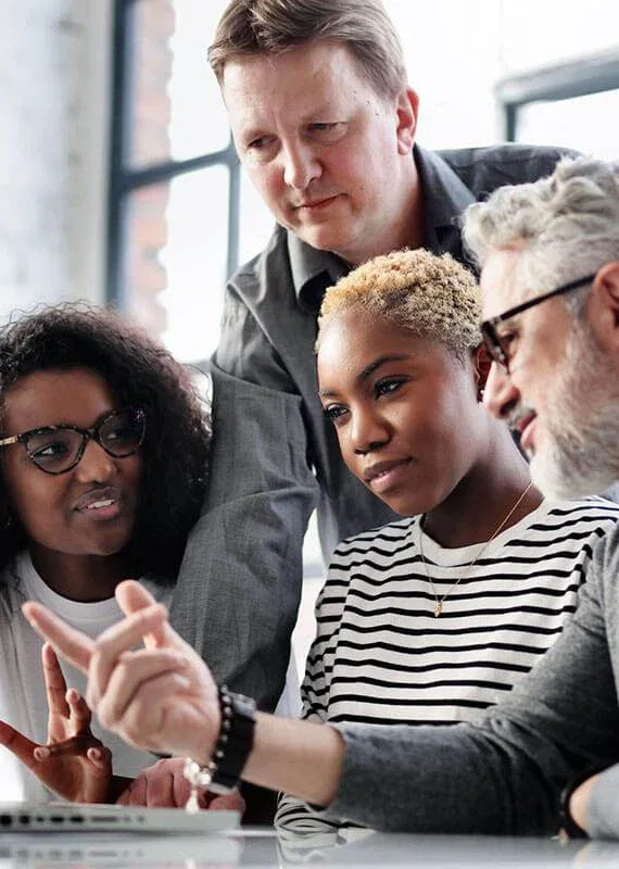 A diverse group of four people collaborate around a laptop, engaged in discussion in a bright office setting.