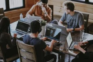 A group of people working on laptops at a wooden table in a casual, collaborative workspace.